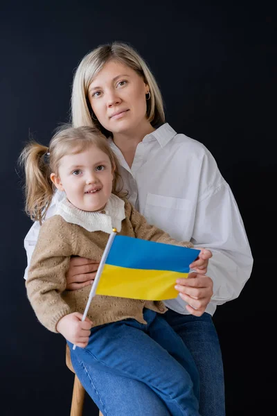 Mujer rubia y niño sonriente con pequeña bandera ucraniana mirando a la cámara aislada en negro - foto de stock