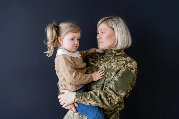 Woman in camouflage holding upset and frowning daughter isolated on black — Stock Photo