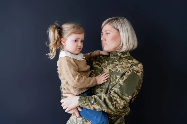 Mujer en camuflaje sosteniendo molesto y frunciendo el ceño hija aislado en negro — Stock Photo