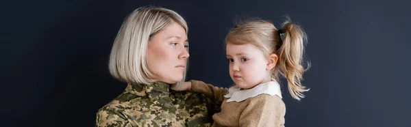 Blonde woman in camouflage looking at upset daughter isolated on black, banner — Stock Photo