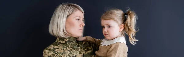 Femme blonde en camouflage regardant fille bouleversée isolé sur noir, bannière — Photo de stock