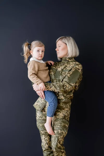 Serious woman in camouflage holding little daughter in hands isolated on black — Stock Photo