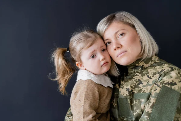 Жінка в камуфляжі дивиться на камеру біля засмученої дочки ізольовано на чорному — стокове фото