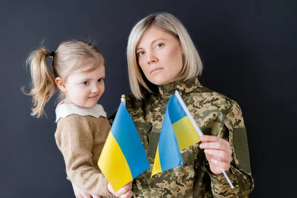 Mujer militar con hija pequeña sosteniendo banderas ucranianas pequeñas aisladas en negro - foto de stock