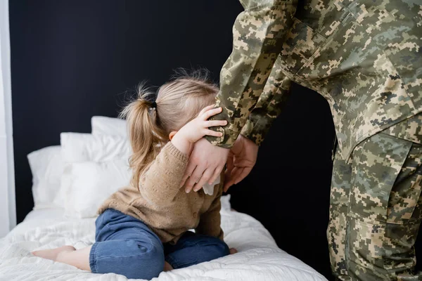 Mujer en uniforme militar cerca de hija pequeña sentada en la cama en casa - foto de stock
