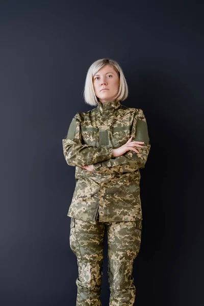 Mujer seria en camuflaje de pie con brazos cruzados aislados en negro — Stock Photo