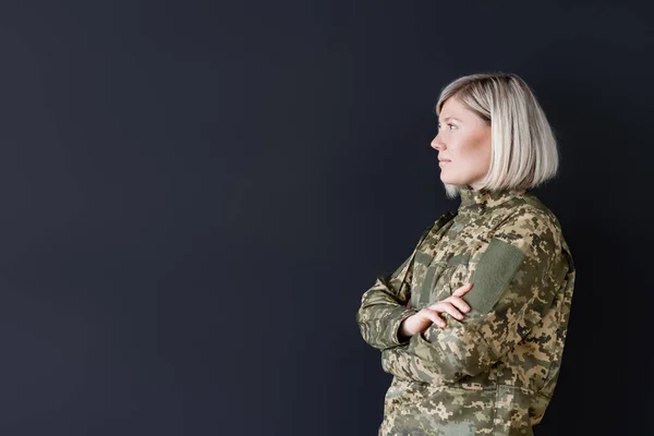 Vista lateral de la mujer en uniforme militar de pie con brazos cruzados aislados en negro — Stock Photo