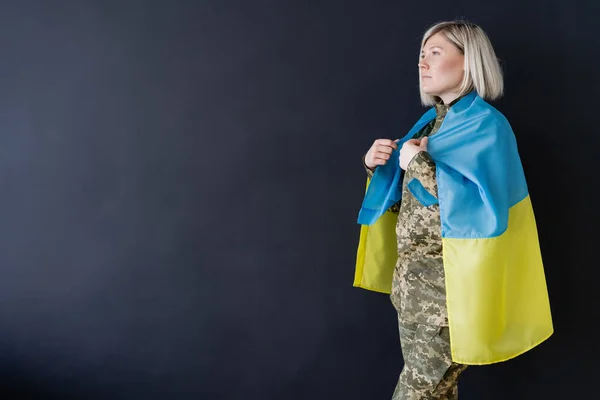 Vista laterale della donna militare con bandiera ucraina sulle spalle guardando lontano isolato sul nero — Foto stock