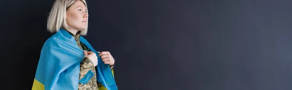 Military woman covered with ukrainian flag looking away isolated on black, banner — Stock Photo