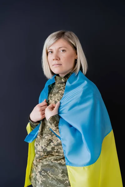 Frau in Militäruniform, mit ukrainischer Flagge auf den Schultern, schaut vereinzelt in die Kamera auf schwarz — Stockfoto