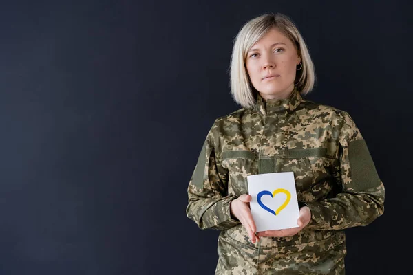 Blonde military woman looking at camera while holding card with blue and yellow heart isolated on black — Stock Photo