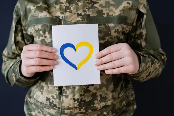 Vue partielle de la femme en uniforme militaire tenant la carte blanche avec le coeur bleu et jaune isolé sur noir — Photo de stock