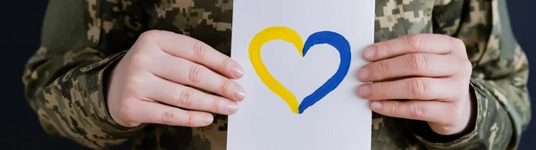 Vista recortada de la mujer militar sosteniendo la tarjeta blanca con corazón azul y amarillo aislado en negro, bandera - foto de stock