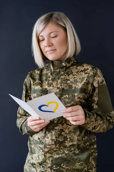 Frau in Militäruniform liest Postkarte mit blauem und gelbem Herz isoliert auf schwarz — Stockfoto