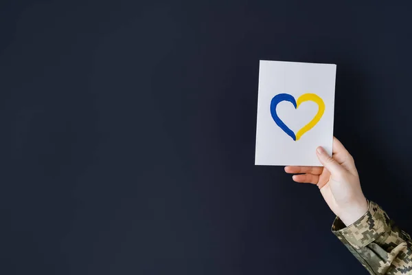 Vista recortada de la mujer en uniforme militar que sostiene la tarjeta blanca con el corazón azul y amarillo aislado en negro — Stock Photo