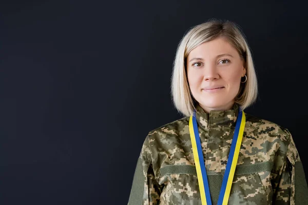 Mujer militar positiva con cinta azul y amarilla mirando a la cámara aislada en negro - foto de stock