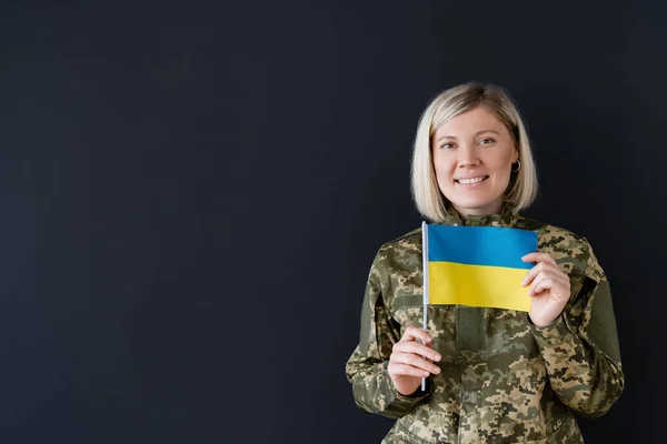 Glücklich blonde Frau in Militäruniform hält kleine ukrainische Flagge isoliert auf schwarz — Stockfoto