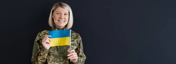 Улыбающаяся женщина в военной форме показывает маленький украинский флаг, изолированный на черном, баннер — стоковое фото