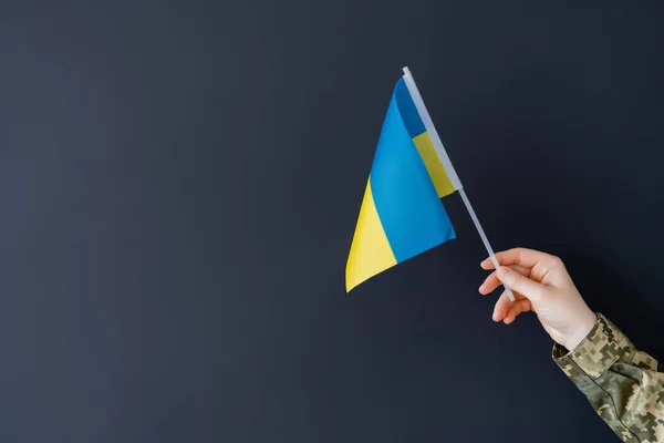 Abgeschnittene Ansicht einer patriotischen Frau in Militäruniform, die eine kleine ukrainische Flagge auf Schwarz hält — Stockfoto