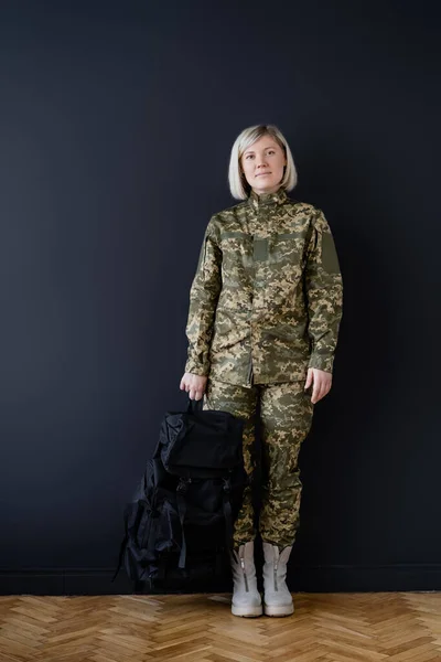 Piena lunghezza della donna in uniforme militare in piedi con zaino nero vicino al muro scuro — Foto stock