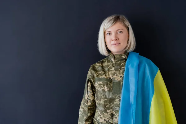 Mujer militar rubia con bandera ucraniana en el hombro mirando la cámara aislada en negro — Stock Photo
