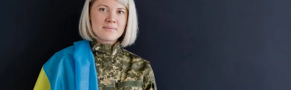 Femme militaire blonde avec drapeau ukrainien regardant la caméra isolée sur noir, bannière — Photo de stock