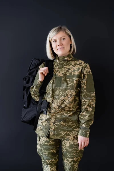 Rubia militar mujer con mochila sonriendo a cámara aislada en negro - foto de stock