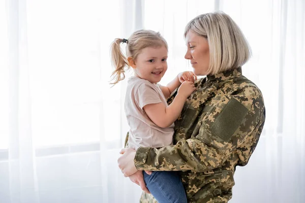 Femme en uniforme militaire tenant fille heureuse à la maison — Photo de stock