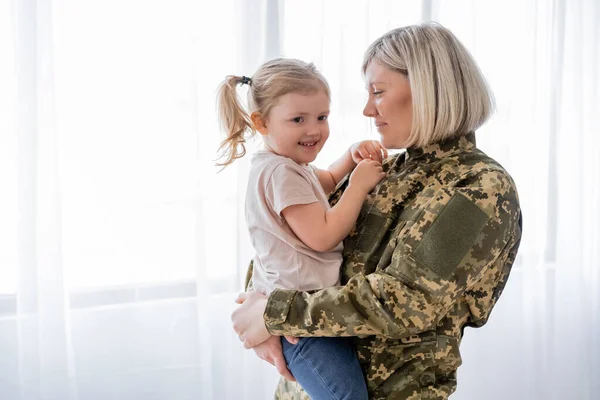 Mujer en uniforme militar sosteniendo feliz hija en casa — Stock Photo
