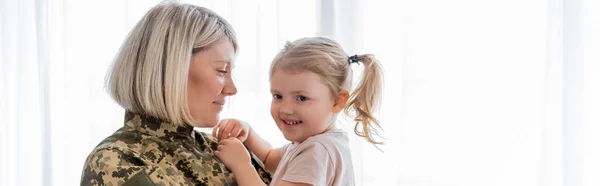 Mujer militar positiva sosteniendo feliz hija en casa, bandera - foto de stock