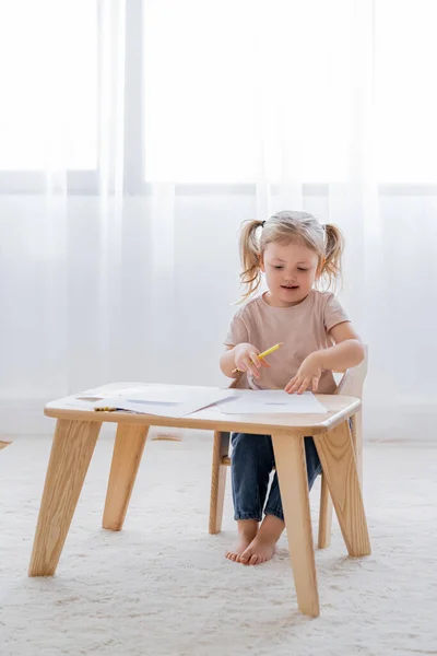 Pleine longueur vue de pieds nus fille tenant crayon de couleur près de papiers sur table en bois — Photo de stock