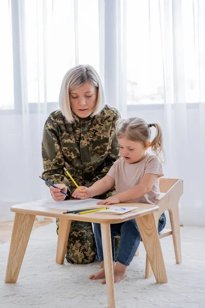 Blonde military woman and little daughter drawing with blue and yellow pencils at home — Stock Photo