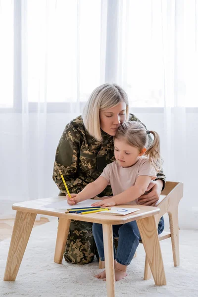 Frau in Militäruniform umarmt kleine Tochter Zeichnung mit blauen und gelben Bleistiften — Stockfoto