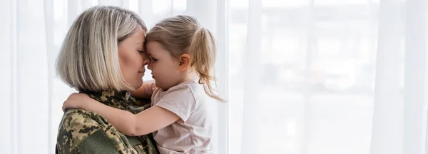 Vista lateral de la mujer militar y la niña abrazando cara a cara cerca de la cortina de ventana blanca, pancarta - foto de stock