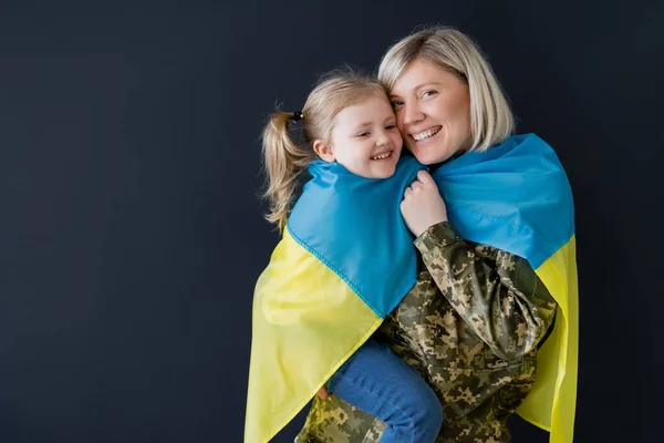 Счастливая военная женщина и дочь, покрытые украинским флагом, обнимающие изолированные на черном — стоковое фото