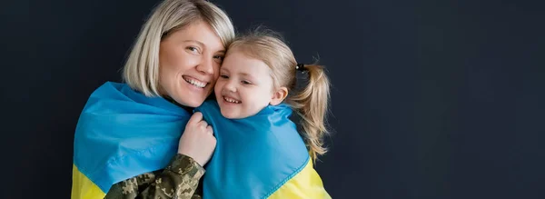 Donna felice guardando la macchina fotografica vicino figlia e bandiera ucraina isolata su nero, banner — Foto stock