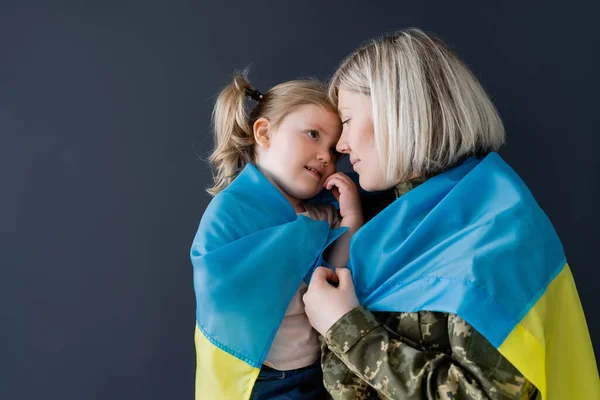 Military woman and little daughter covered with ukrainian flag looking at each other isolated on black — Stock Photo