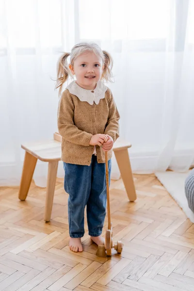 Vista completa de la chica descalza con juguete de madera sonriendo a la cámara - foto de stock