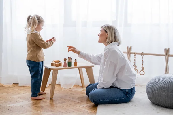 Side view of woman pointing with hand near daughter playing with wooden figurines at home — Stock Photo