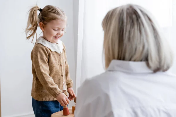 Allegra ragazza che gioca con figurine di legno vicino alla madre offuscata — Foto stock