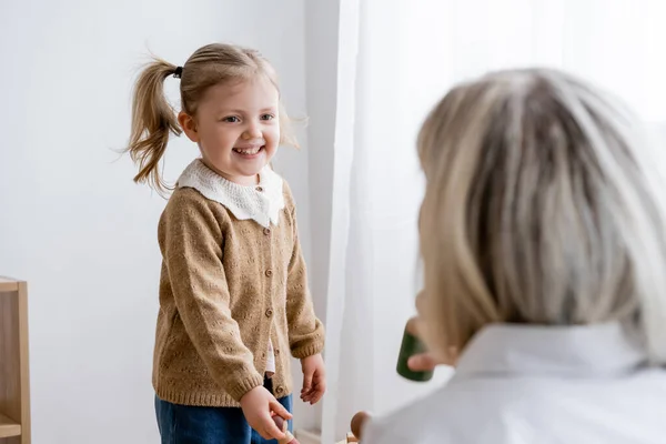 Cheerful girl with ponytail smiling near blurred mom at home — Stock Photo