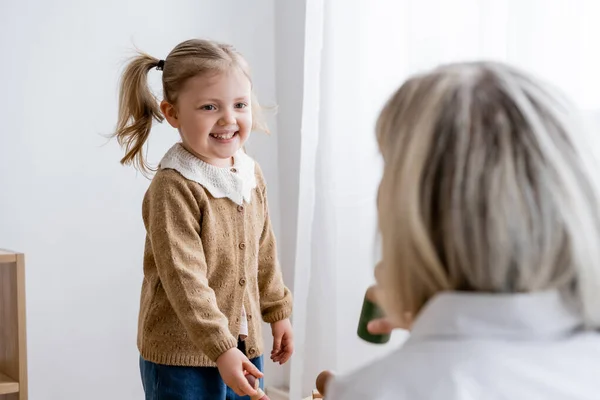 Fille gaie avec queue de cheval souriant près de maman floue à la maison — Photo de stock