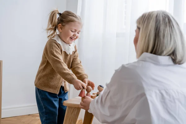 Bambino ridente che gioca con figurine di legno vicino alla mamma offuscata a casa — Foto stock