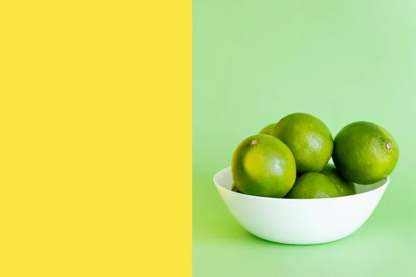 Limes maduros em tigela sobre fundo verde e amarelo — Fotografia de Stock