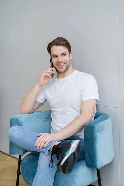 Fröhlicher Mann sitzt im Sessel neben grauer Wand und spricht auf Smartphone — Stockfoto