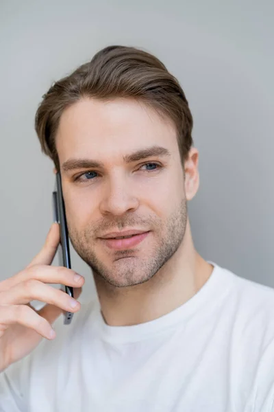 Retrato del joven mirando hacia otro lado durante la conversación en el teléfono inteligente aislado en gris - foto de stock