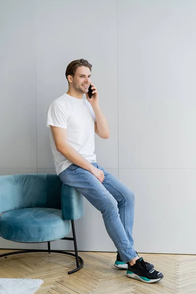 Visão de comprimento total do homem sorridente em jeans falando no smartphone perto de poltrona e parede cinza — Fotografia de Stock