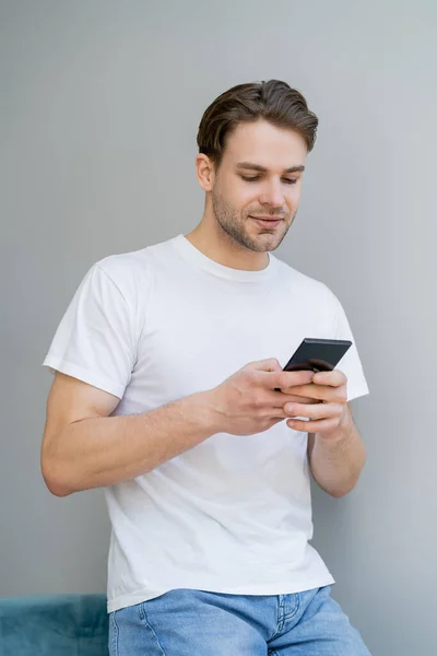 Jeune homme positif en t-shirt blanc bavardant sur smartphone sur fond gris — Photo de stock