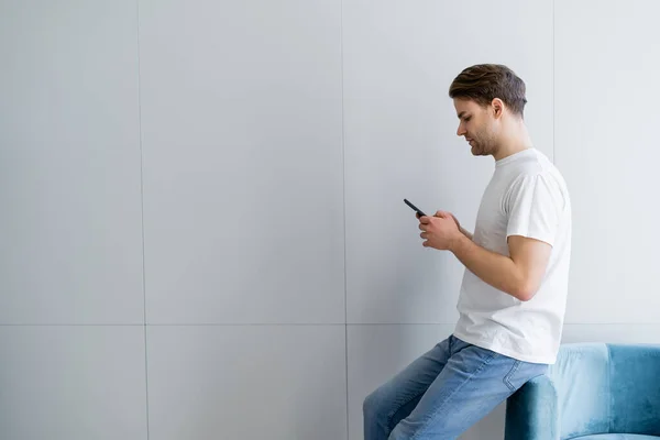 Seitenansicht eines jungen Mannes im weißen T-Shirt mit Handy in der Nähe einer grauen Wand — Stockfoto