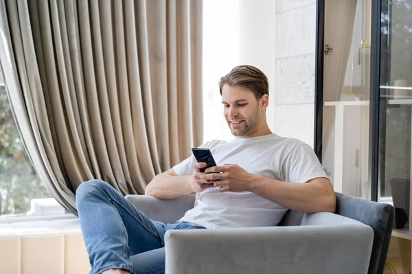 Задоволений чоловік у білій футболці обмін повідомленнями на мобільний телефон, сидячи в кріслі — стокове фото